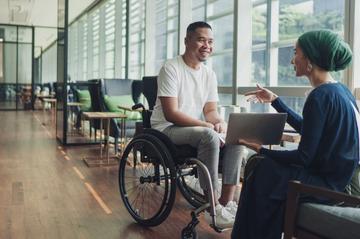 Person using a wheelchair having a conversation with another person holding a laptop in a modern office.