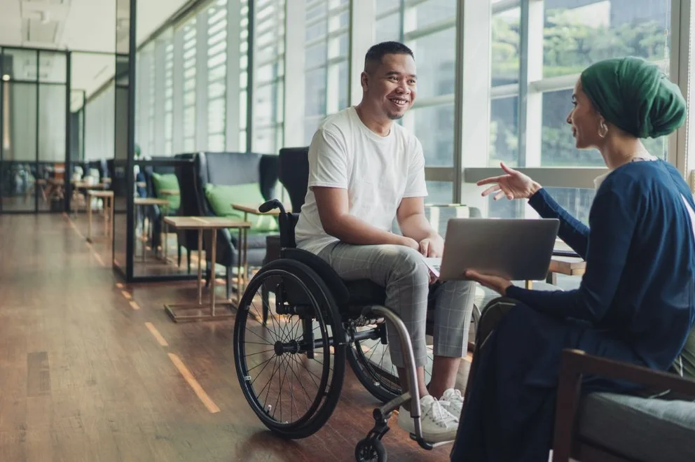 Person using a wheelchair having a conversation with another person holding a laptop in a modern office.