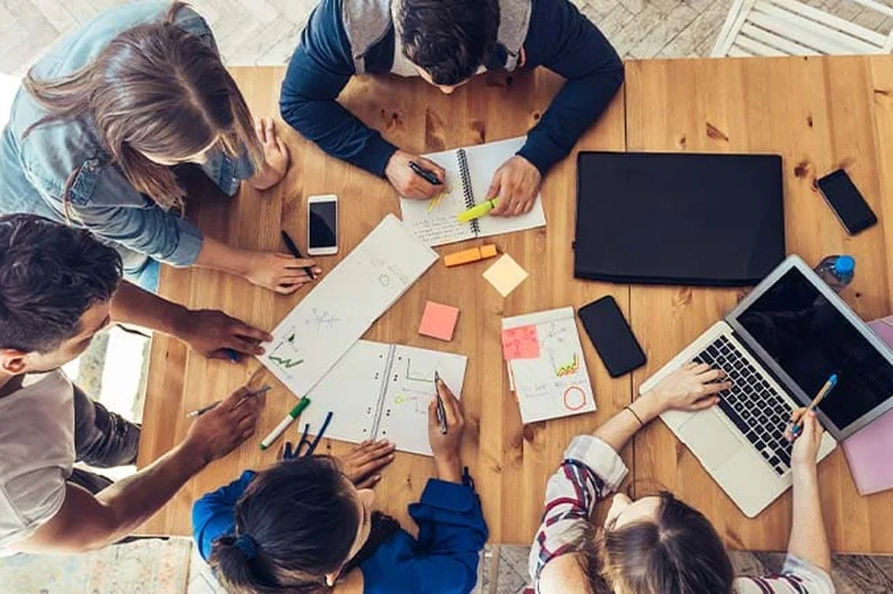 An overhead view of a group of people sat around a wooden table that is filled with paper, notepads, a laptop and a tablet.