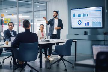 A group of people are in an office meeting room. A man is stood at the front of the room in front of a screen, presenting to the others who are sat around the table.