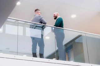 Professional setting with two people standing on an indoor glass balcony, engaged in conversation within a modern office building with overhead lighting.