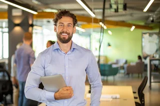 Person holding a laptop and standing in a modern open-plan office, with colleagues and workspaces visible in the background.