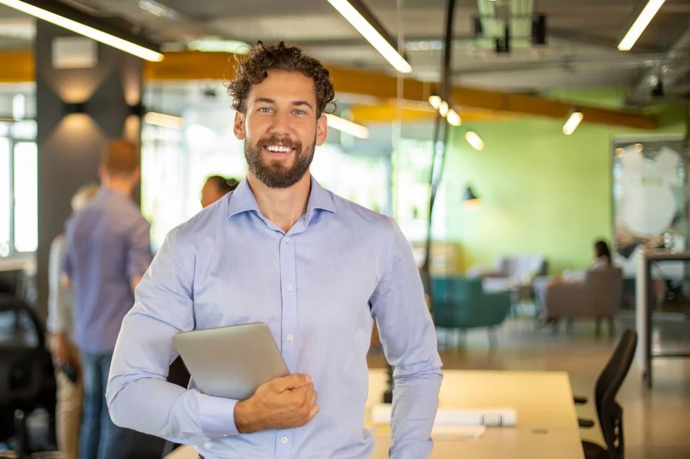 Person holding a laptop and standing in a modern open-plan office, with colleagues and workspaces visible in the background.