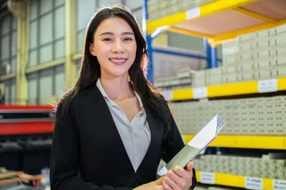 Person holding documents while standing in a warehouse aisle, with industrial shelving and stacked materials visible in the background.