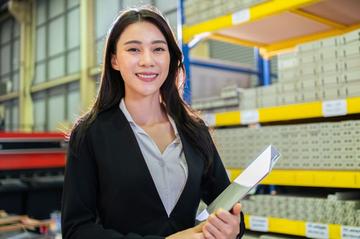 Person holding documents while standing in a warehouse aisle, with industrial shelving and stacked materials visible in the background.