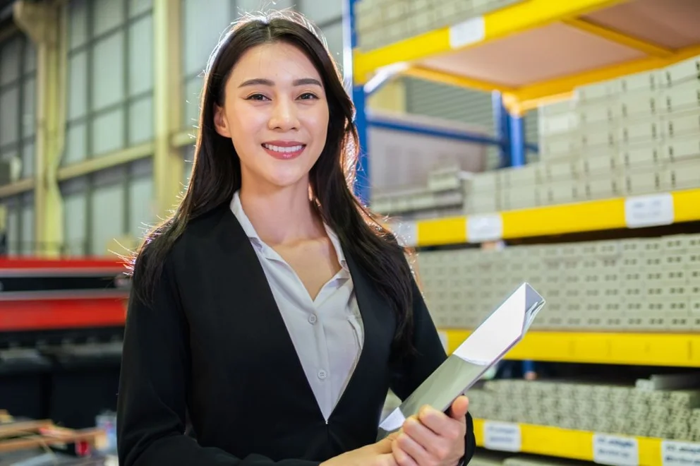 Person holding documents while standing in a warehouse aisle, with industrial shelving and stacked materials visible in the background.
