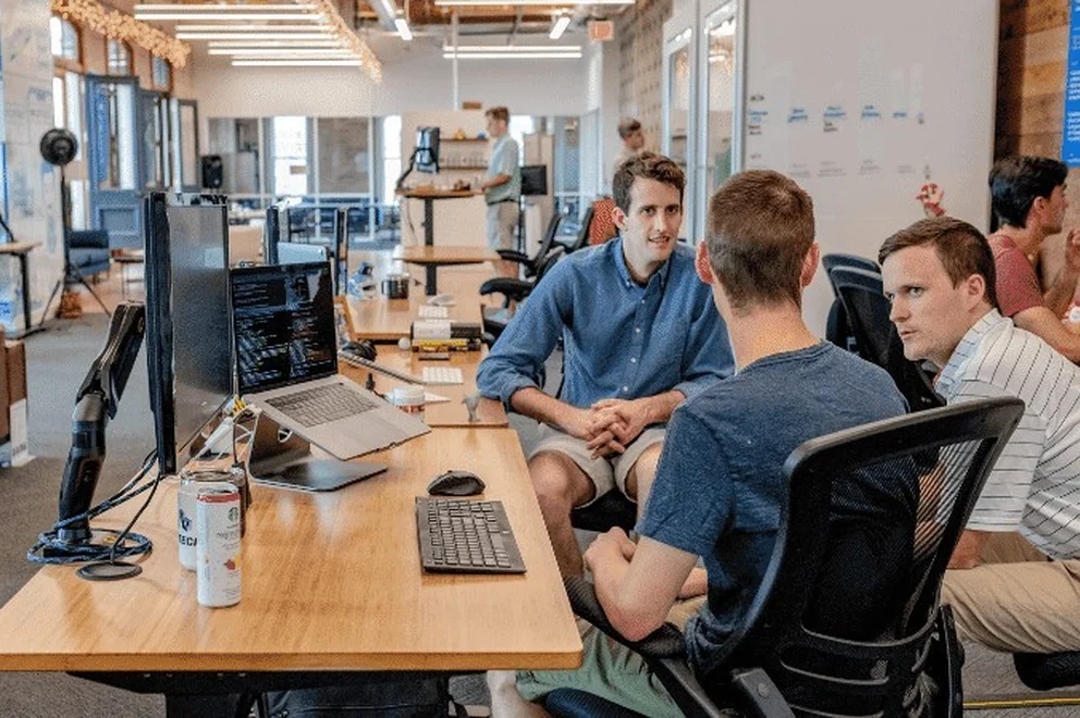 Three men sitting in office chairs and chatting at a desk with computer screens on their left