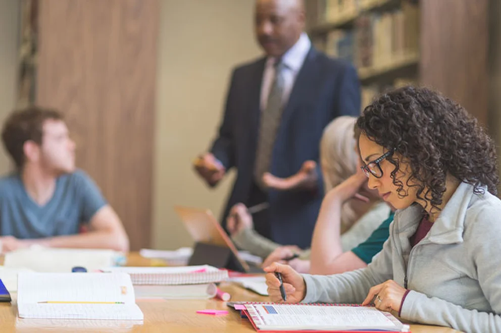 A woman sits at a desk in a group education setting while taking notes.