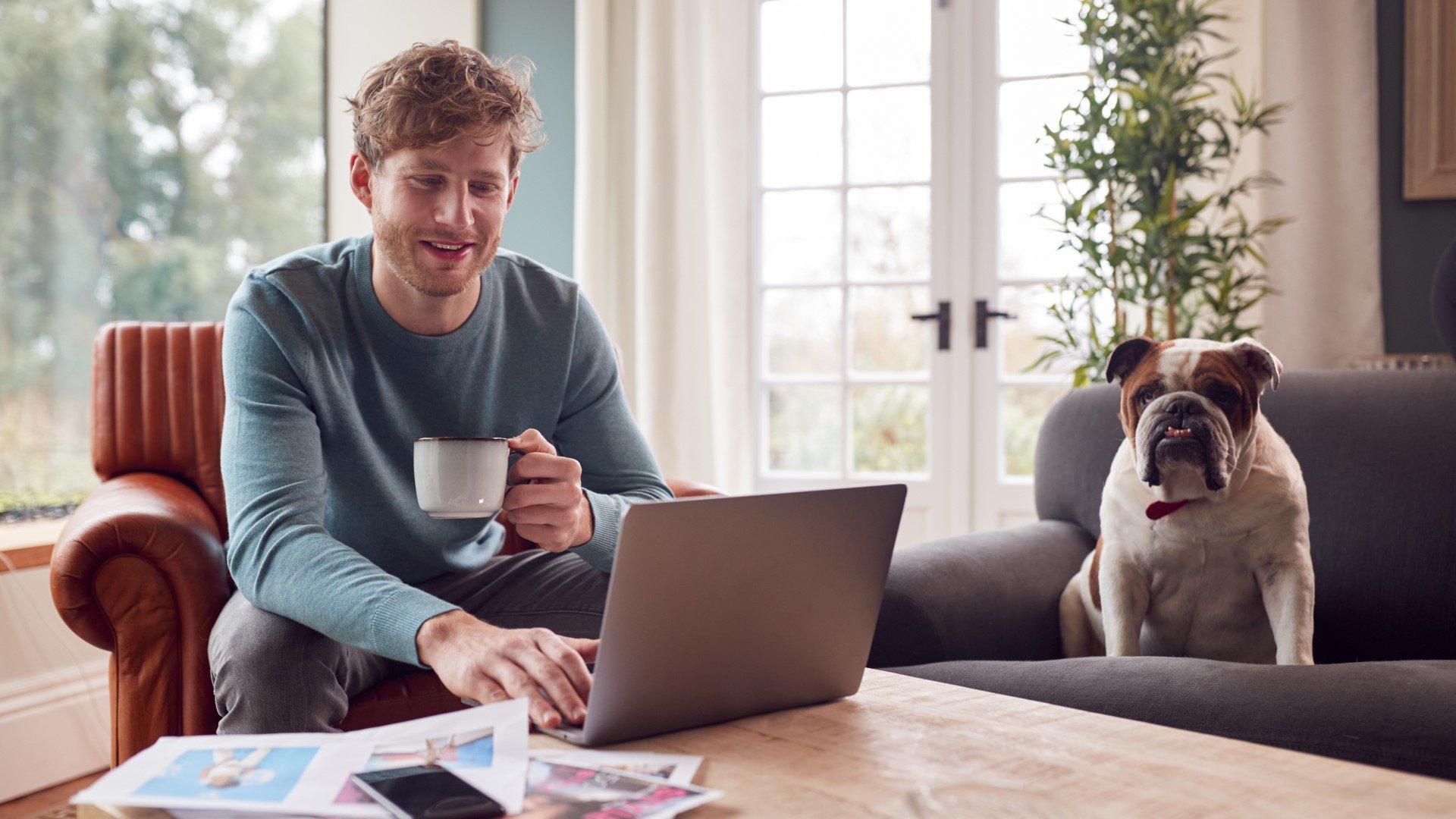 man-holding-coffee-on-laptop-with-bull-dog-on-sofa