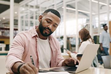 A man is working or studying using a laptop and notepad, He's sat at a desk in a public place.