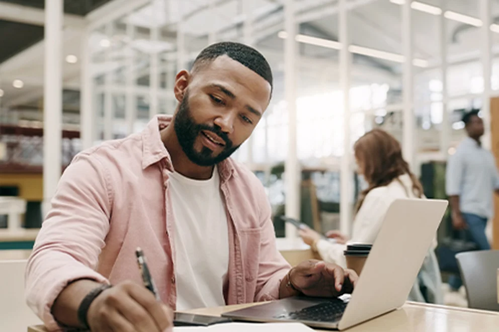 A man is working or studying using a laptop and notepad, He's sat at a desk in a public place.