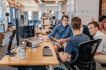 Three men sitting in office chairs and chatting at a desk with computer screens on their left