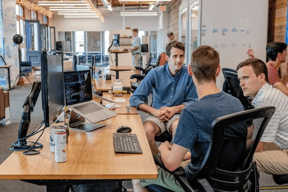 Three men sitting in office chairs and chatting at a desk with computer screens on their left