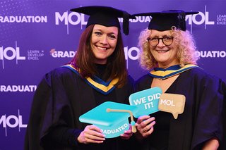 Two MOL learners in cap and gowns pose with signs that say 'We did it' at the MOL graduation