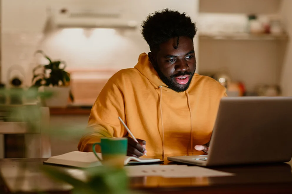 A man is sat at the table at home. He's on his laptop with a brew and study materials on the table.