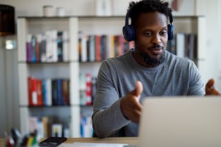 A man is sat at a desk and wearing headphones while he talks to someone on his laptop.