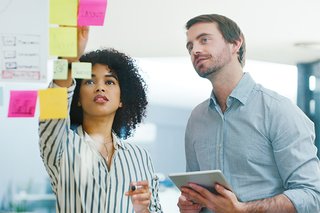 A man and woman are standing in front of a glass screen, looking at the sticky notes on there.