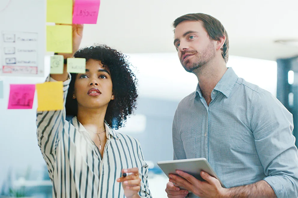 A man and woman are standing in front of a glass screen, looking at the sticky notes on there.