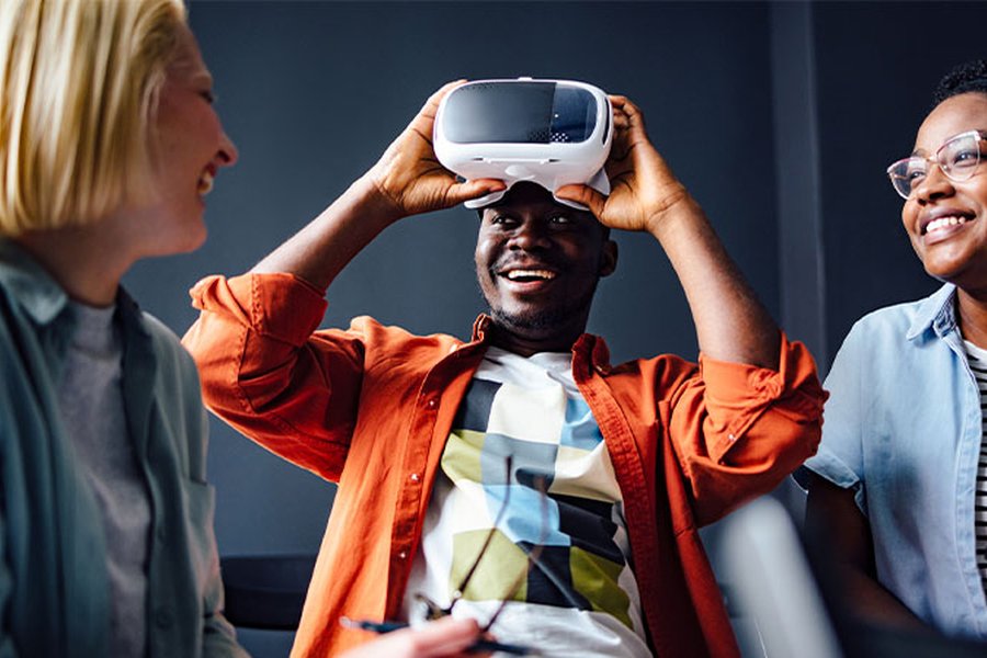 A man is smiling as he takes off a VR headset. Two women are next to him on either side and they are laughing.