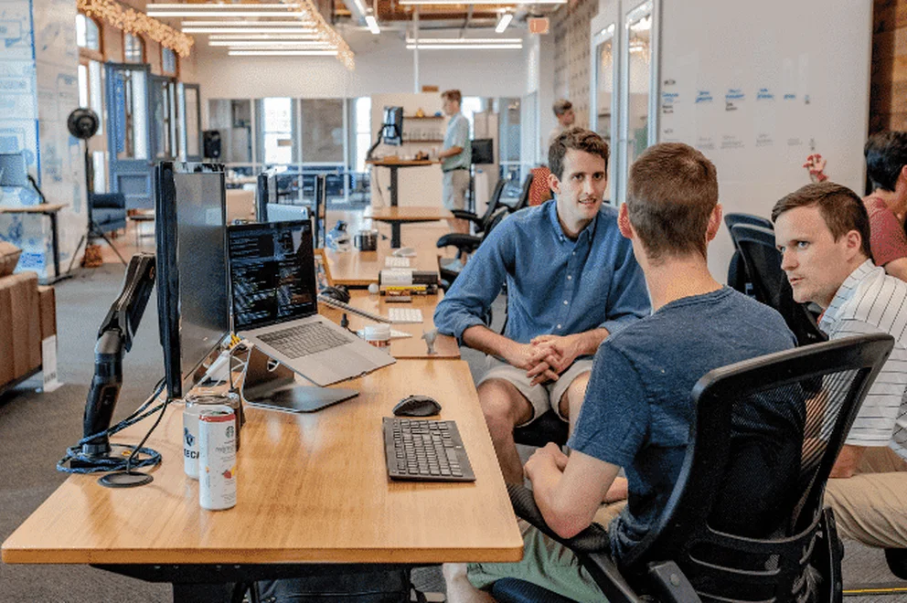 Three men sitting in office chairs and chatting at a desk with computer screens on their left