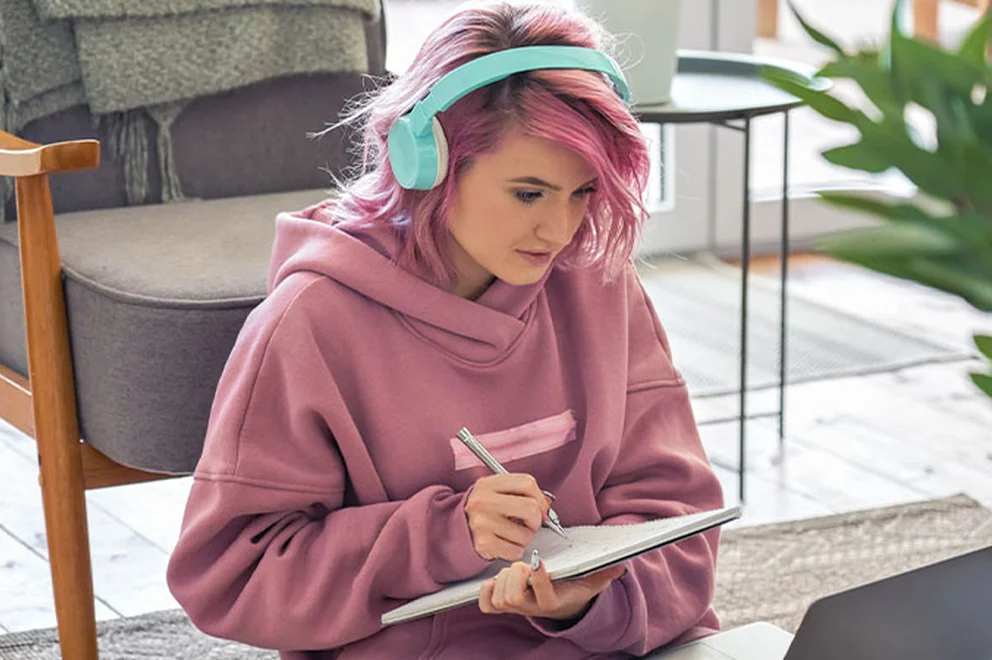 A young woman is sat on the floor, watching something on her laptop and taking notes.