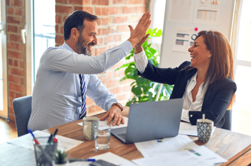Two people giving each other high five sitting at a desk with laptop computers