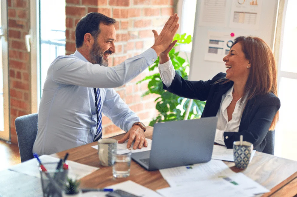 Two people giving each other high five sitting at a desk with laptop computers