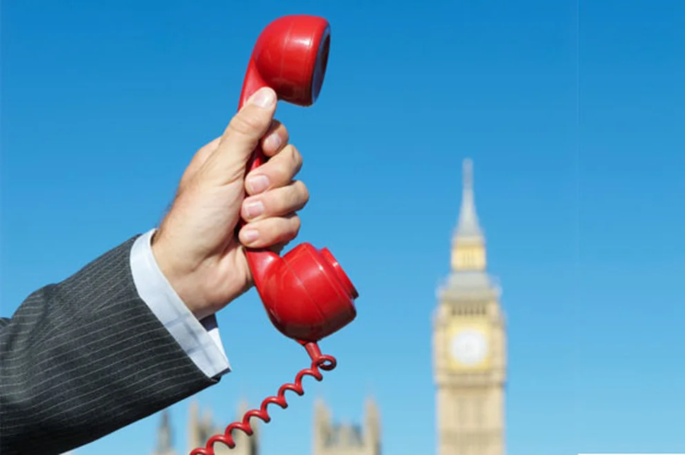 Close up of the hand of a man wearing a suit and holding a red telephone receiver with British parliament on the background