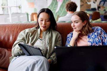 Two women are sat working together in a cafe, one with a tablet and one with a laptop.