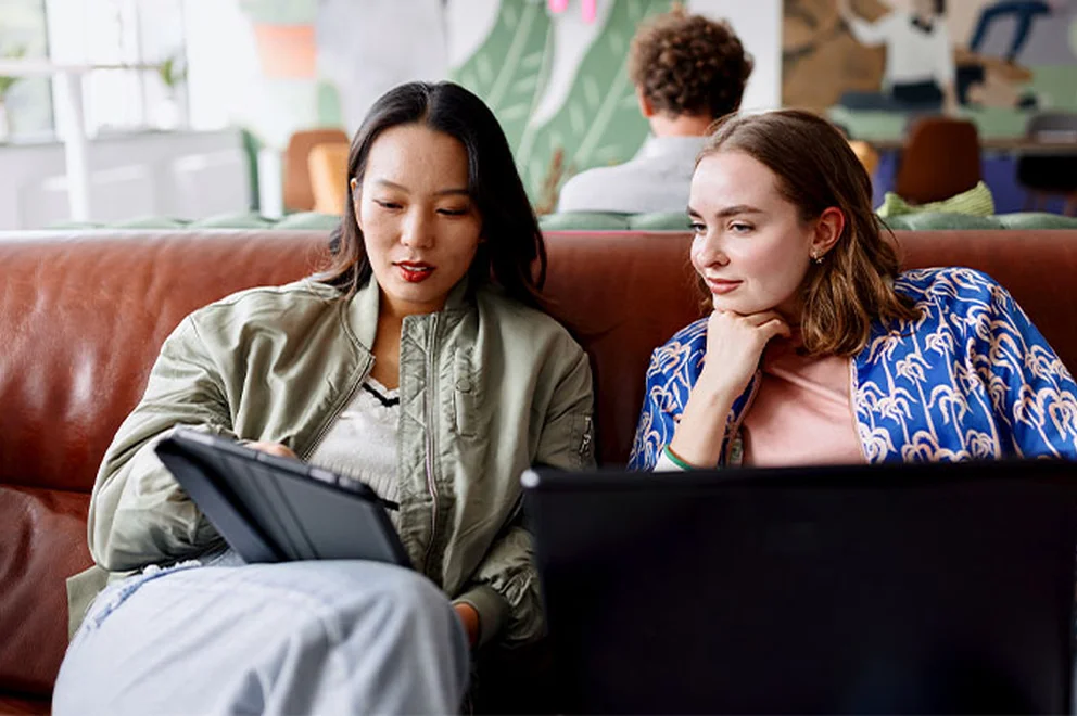 Two women are sat working together in a cafe, one with a tablet and one with a laptop.