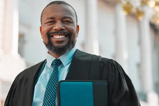 A man in a shirt, tie and black robe is standing in front of a courthouse and holding a folder. He's smiling at the camera.