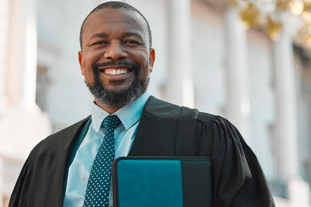 A man in a shirt, tie and black robe is standing in front of a courthouse and holding a folder. He's smiling at the camera.