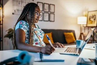 A lady is sat at her desk at home. We have a side-on view as she looks at her laptop screen and takes notes.