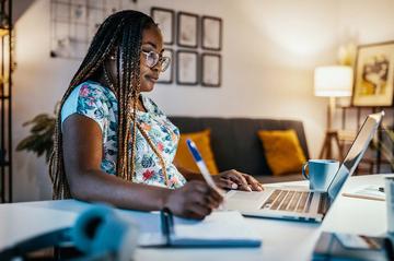 A lady is sat at her desk at home. We have a side-on view as she looks at her laptop screen and takes notes.