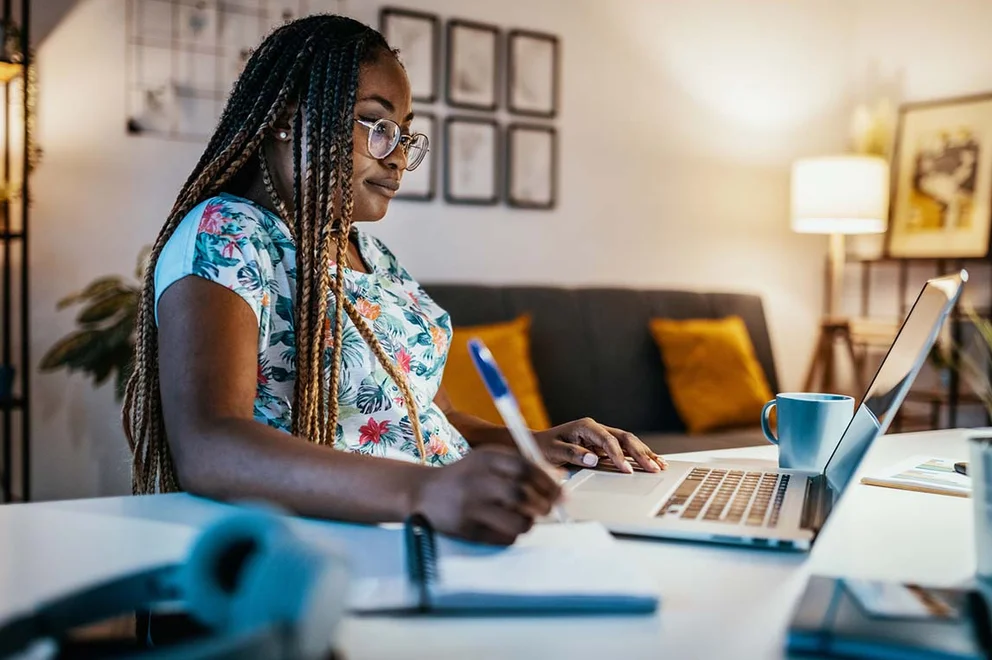 A lady is sat at her desk at home. We have a side-on view as she looks at her laptop screen and takes notes.