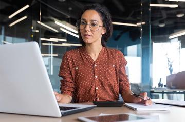 A woman is sat at a desk in an office. She is looking at a laptop and while taking notes.