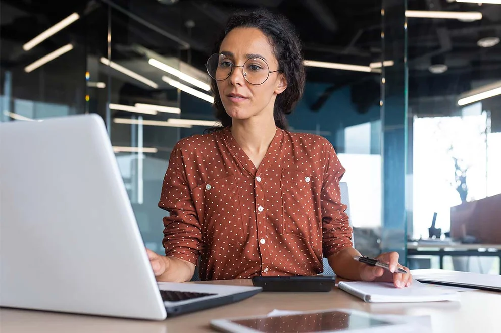 A woman is sat at a desk in an office. She is looking at a laptop and while taking notes.