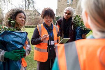 A group of people are working outdooers in orange hi-viz jackets. They are stood around their manager who looks like they are giving out instructions.