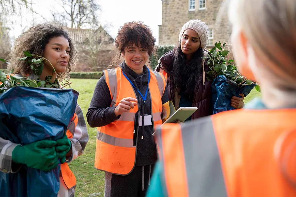 A group of people are working outdooers in orange hi-viz jackets. They are stood around their manager who looks like they are giving out instructions.
