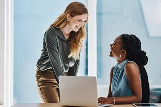 Two professionals collaborating at a desk with a laptop in a modern office, discussing work and reviewing HR statistics together.