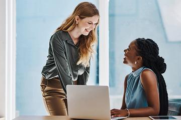 Two professionals collaborating at a desk with a laptop in a modern office, discussing work and reviewing HR statistics together.