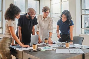 A group of workers are in an office, standing around a desk with lots of papers on it.
