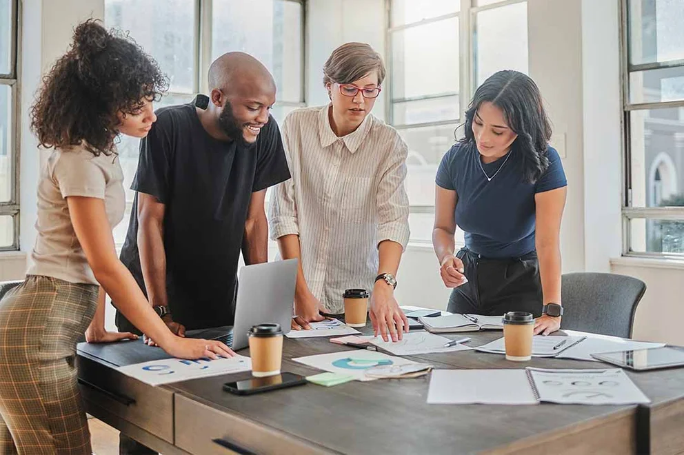 A group of workers are in an office, standing around a desk with lots of papers on it.