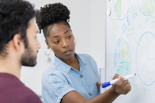 A lady is drawing charts on a whiteboard with a male colleague.