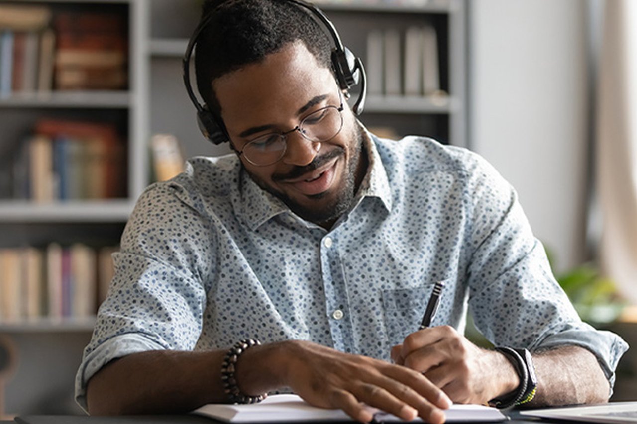 A man is taking notes while sat at his laptop wearing headphones.