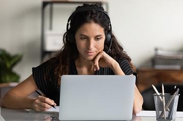 A woman is sat at a desk wearing headphones and looking at her laptop screen. She is taking notes.