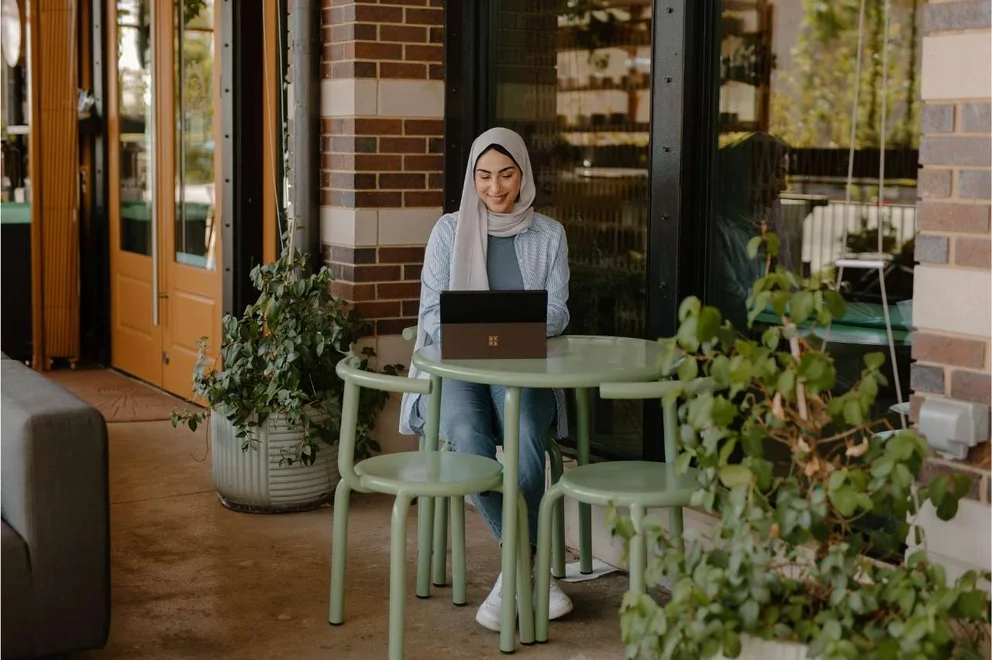 Young person working on a laptop computer while stting at a green table outside a building surrounded by plants